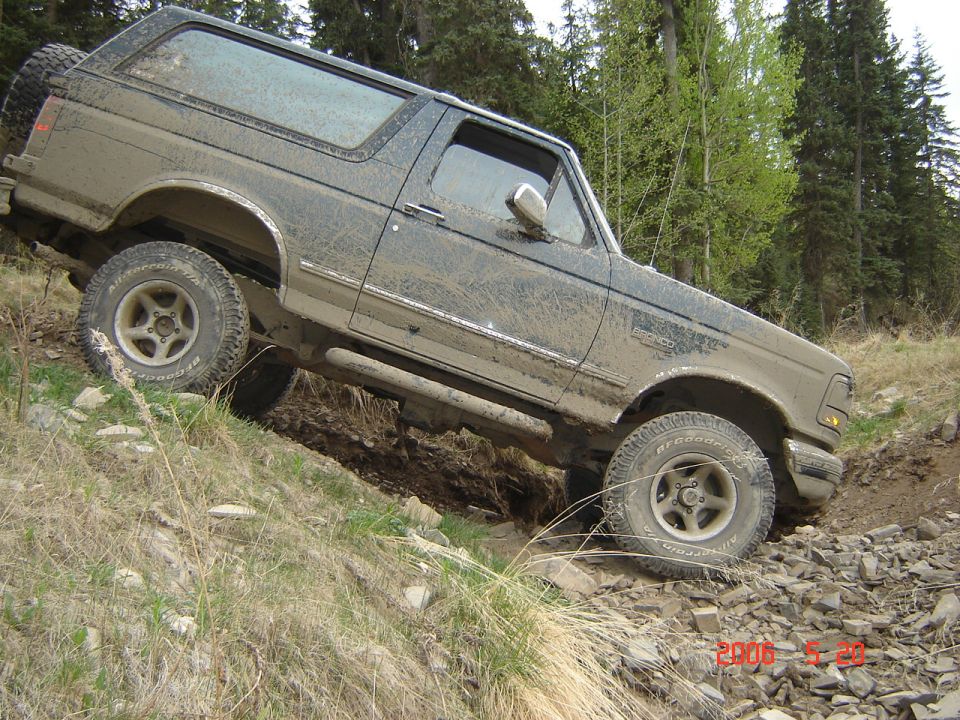 Mud-covered dark green 1992 Ford Bronco driving off-road on a rocky incline