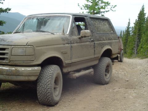 1995 Ford Bronco - This 1995 Ford Bronco is seen off-road, covered in mud on a dirt path with trees in the background.