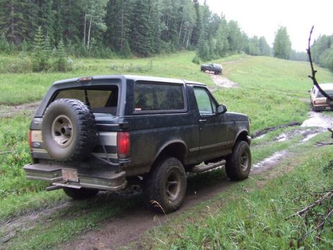 1995 Ford Bronco - A black 1995 Ford Bronco navigates a muddy off-road path through a wooded area.
