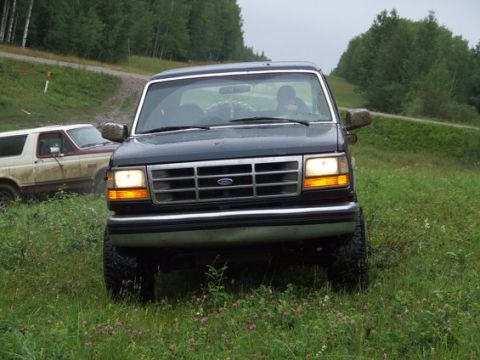 1995 Ford Bronco - Two Ford Broncos are parked off-road in a grassy area with trees in the background.