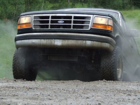 1995 Ford Bronco - This black Ford Bronco is captured from the front as it drives on a dirt road, creating dust clouds.