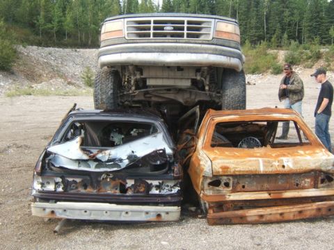 1995 Ford Bronco - A large Ford truck is parked on top of two heavily damaged cars in a junkyard setting.