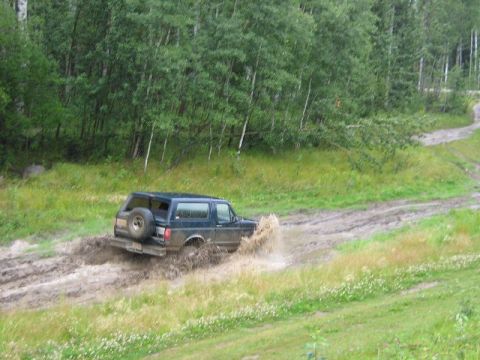 1995 Ford Bronco - A dark 1995 Ford Bronco navigates a muddy off-road trail, kicking up water and mud.