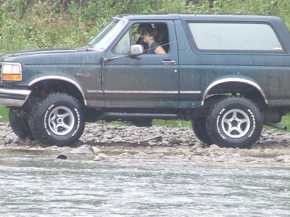 Green 1995 Ford Bronco driving through shallow water on a rocky shore