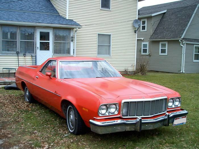 Red 1970s Ford Ranchero parked on grass beside a house, front three-quarter view.
