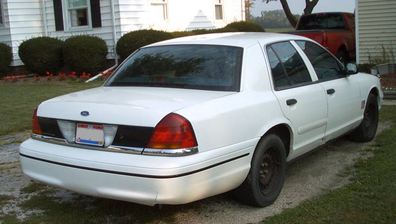 Rear view of a white 1999 Ford Crown Victoria with black trim and a red truck in the background