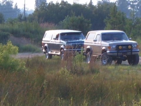 1993 Ford Bronco - Two Ford Broncos, one dark green with a camper shell and the other tan with off-road lights, are parked on a dirt road.