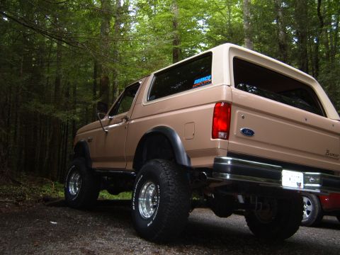 1993 Ford Bronco - Rear view of a lifted tan 1993 Ford Bronco with aftermarket wheels in a wooded area.
