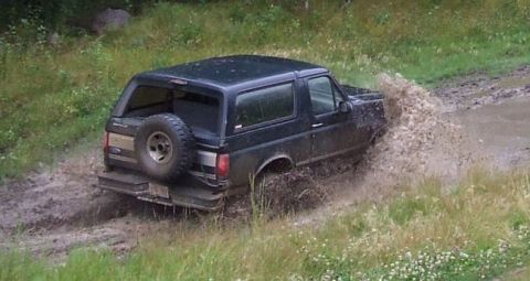 1988 Ford Bronco - A dark SUV navigates a muddy track, kicking up a large spray of water.