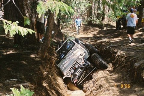 1966 Jeep CJ5 - A Jeep is stuck in a deep rut on a wooded trail, with onlookers present and another vehicle nearby.