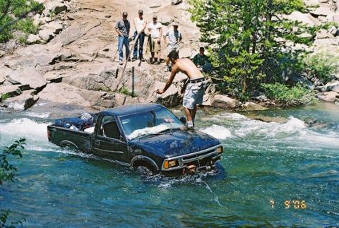 1966 Jeep CJ5 - A black pickup truck navigates a rocky riverbed while onlookers observe from the bank.