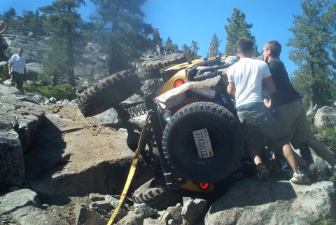 1966 Jeep CJ5 - A yellow Jeep Wrangler is stuck on its side on a rocky off-road trail, with two people attempting to push it upright.