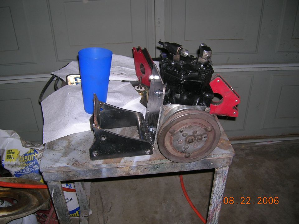 Black air conditioning compressor with red brackets and pulley, on a metal table next to a blue cup