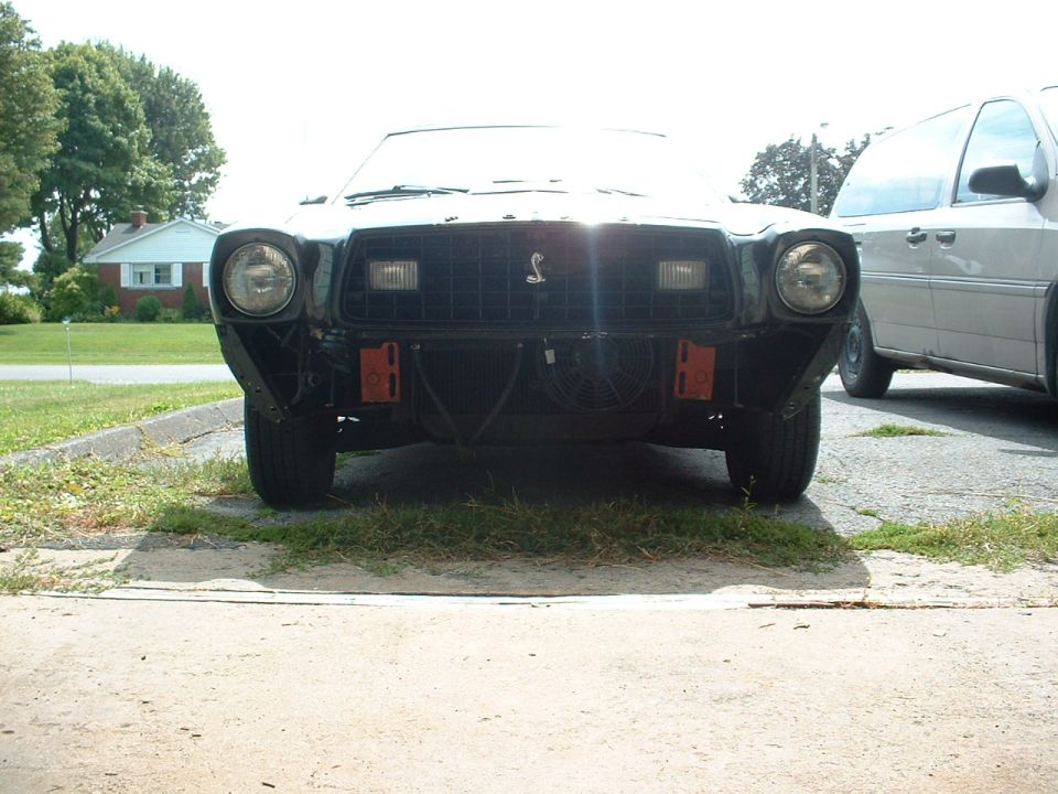1978 Ford Mustang II front end, missing bumper, with a silver van parked to the right