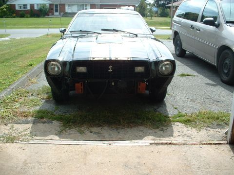 1978 Ford Mustang II - A black 1978 Ford Mustang II is parked in front of a garage, with a minivan to its right.