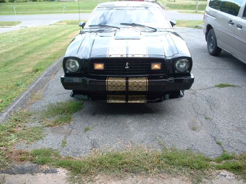 1978 Ford Mustang II - A black 1978 Ford Mustang II Cobra II with gold racing stripes is parked in a driveway next to a silver van.