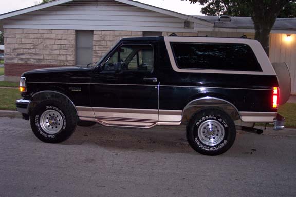 Black two-tone Ford Bronco, side profile, parked at curb in front of brick house at dusk