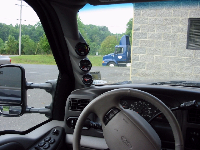 Interior with three aftermarket gauges on A-pillar pod, gray Ford steering wheel and dash, towing mirror visible
