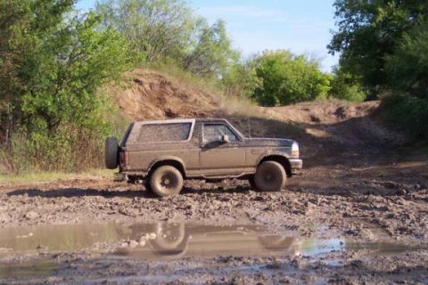 1996 Ford Bronco - A 1996 Ford Bronco navigates a muddy off-road trail, kicking up water as it drives through a large puddle.