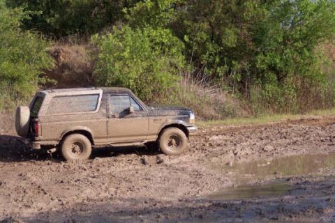 1996 Ford Bronco - A muddy Ford Bronco navigates a challenging off-road terrain with deep mud puddles.