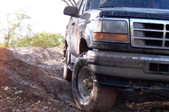 Mud-covered black Ford Bronco on a dirt track, front 3/4 view