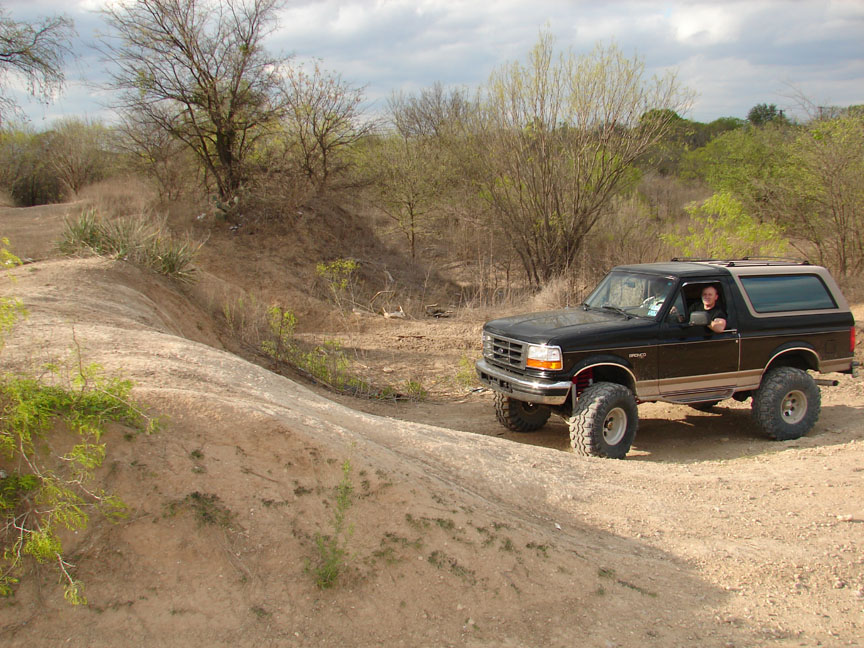Lifted black and tan Ford Bronco on big tires parked on a dirt off-road trail beside a gully