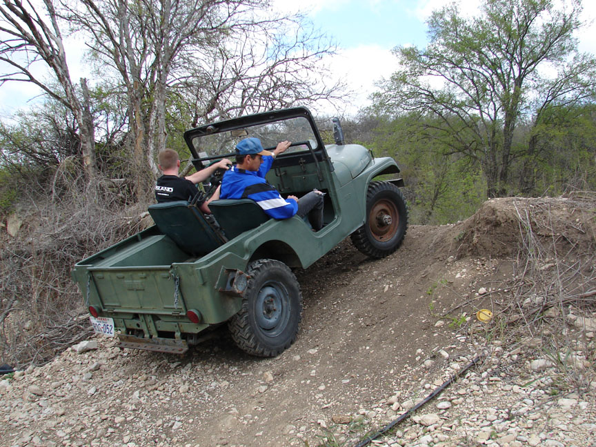 Two men in an olive-drab military flatbed Jeep climbing a rocky dirt embankment off-road