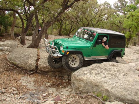1996 Ford Bronco - A Jeep Wrangler navigates a rocky trail, with a person visible in the trees in the background.