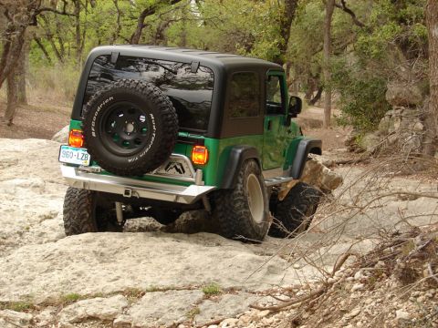 1996 Ford Bronco - A green Jeep Wrangler is shown off-roading on a rocky path, with its spare tire mounted on the back.