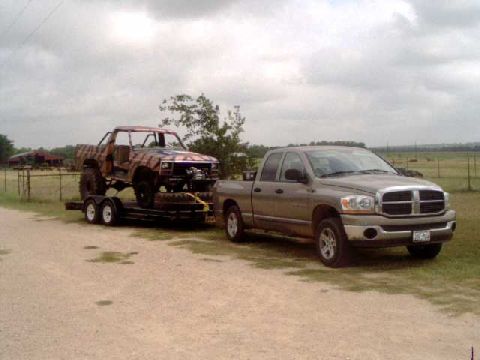 1984 Ford Bronco - A pickup truck tows a custom-built off-road vehicle on a trailer under a cloudy sky.