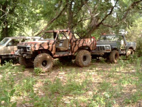 1984 Ford Bronco - Two off-road vehicles, a camouflaged Bronco and a dark pickup, are parked in a wooded area.