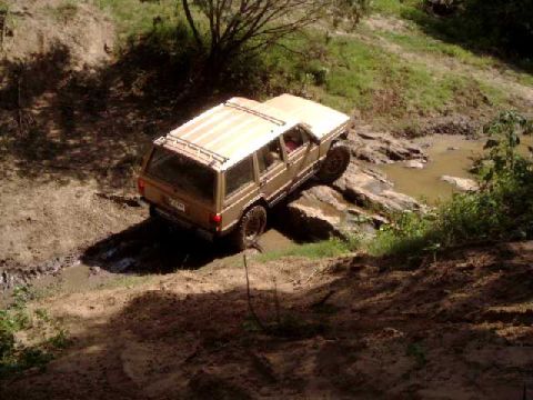 1984 Ford Bronco - A tan Jeep Cherokee XJ drives through a rocky stream bed, kicking up mud.