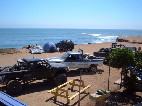 1985 Ford Ranger - Multiple off-road trucks and camping gear are visible on a beach next to the ocean.