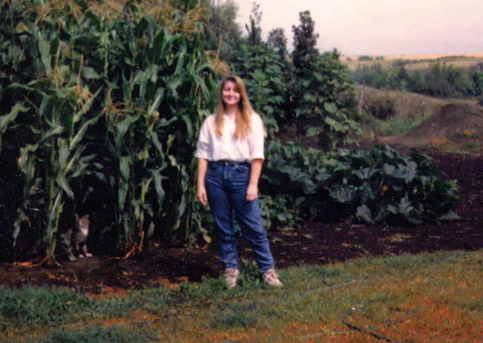 Young woman in white shirt and jeans standing on grass beside tall corn and a garden, cat nearby