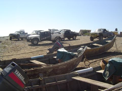 1985 Ford Ranger - A collection of modified pickup trucks and weathered fishing boats are staged on a sandy shoreline under a clear blue sky.