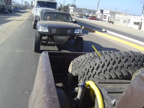 1985 Ford Ranger - A modified pickup truck tows a trailer down a paved road with buildings lining the side.