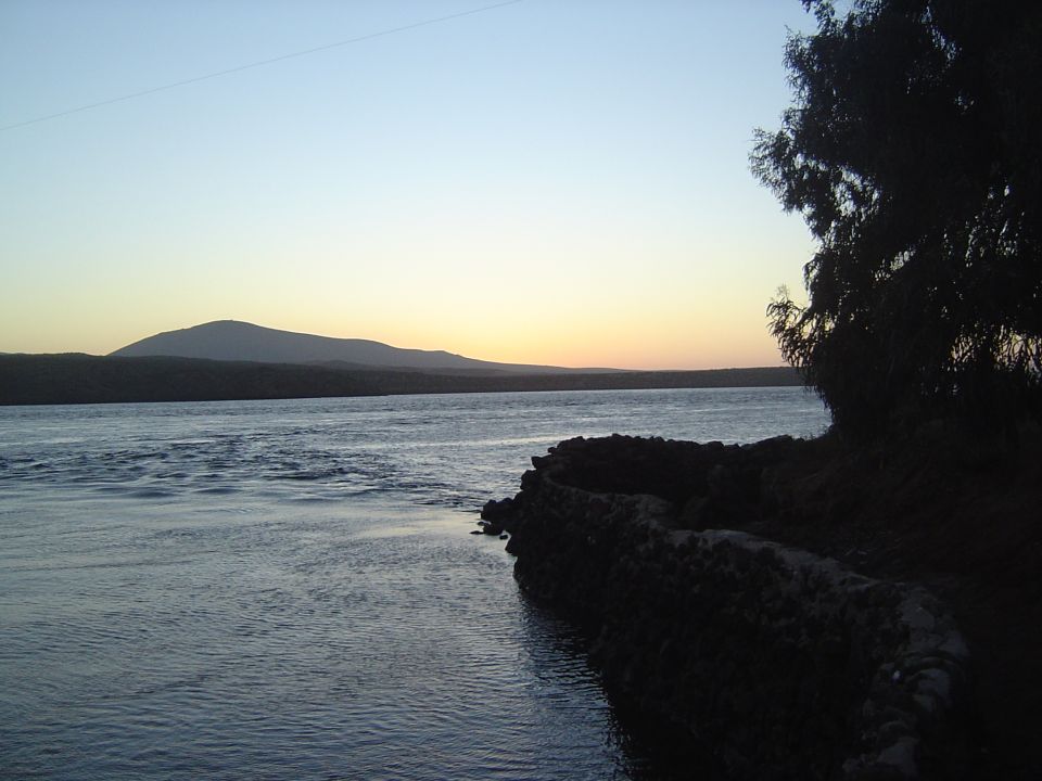 River flows past a rocky shore with a tree silhouette against a sunset sky and distant mountains