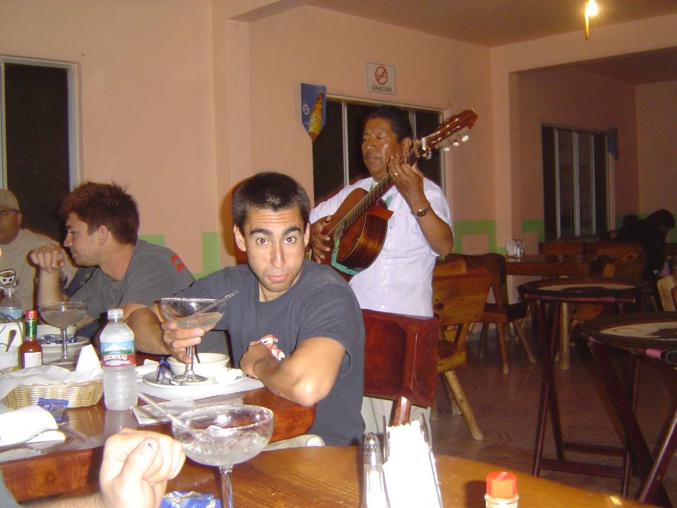 guy holding a margarita looks at the camera while a musician plays guitar behind him