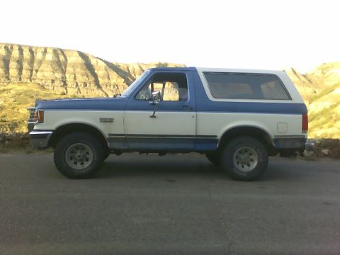 1989 Ford Bronco - A two-tone blue and white Ford Bronco is seen from the side, parked on a road with a natural landscape behind it.