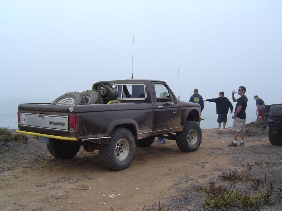 Brown Ford Ranger pickup truck parked on a dirt cliff overlooking the ocean, spare tires in the bed