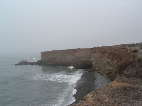 1985 Ford Ranger - Waves break against a pebble beach under a large cliff on a foggy day.