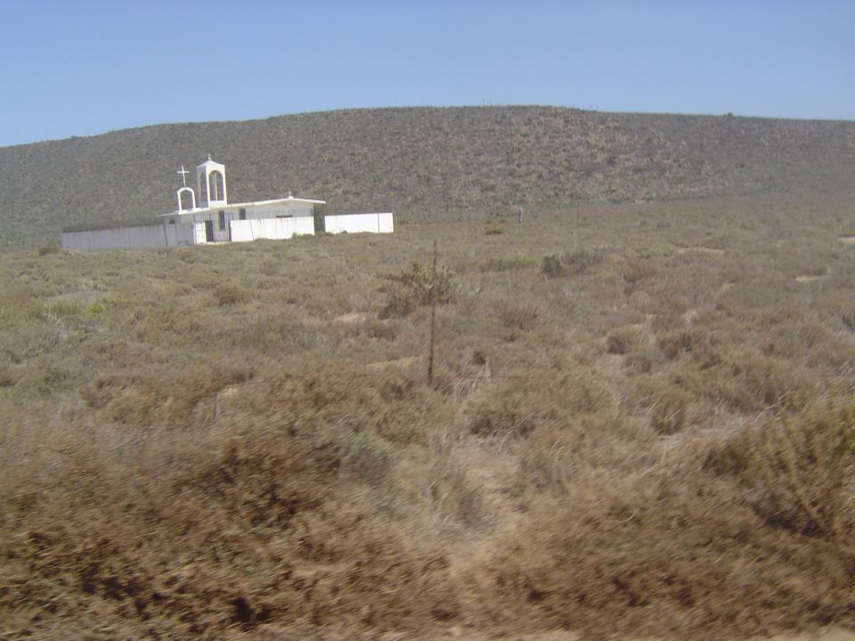 White church building with a bell tower sits on a hill in a dry, scrubby landscape under a clear blue sky