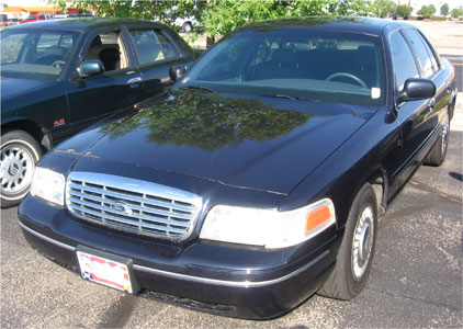 1999 Ford Crown Victoria - A black Ford Crown Victoria is parked next to another sedan, with trees and a building in the background.