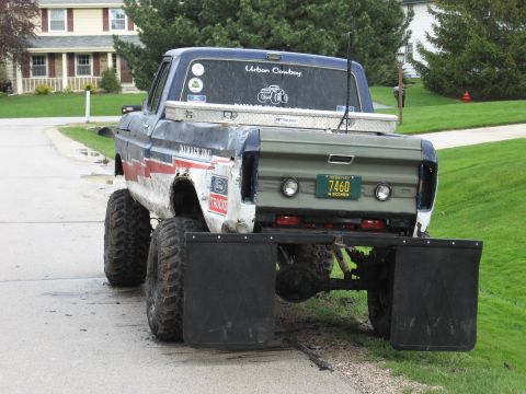 1977 Ford F-150 - Rear view of a lifted vintage Ford pickup on a residential street, wearing weathered white, red and blue 'Damage Inc' graphics with an oliv…
