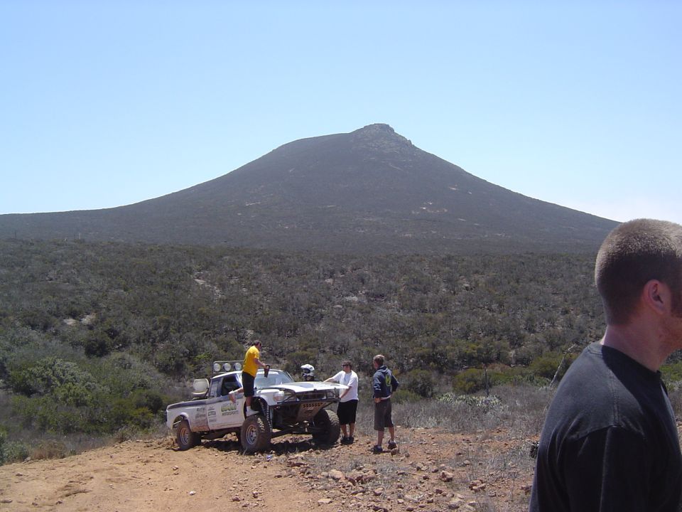 White off-road pickup truck with large tires, modified front end, and people gathered around, parked on a dirt track with a mountain in the background