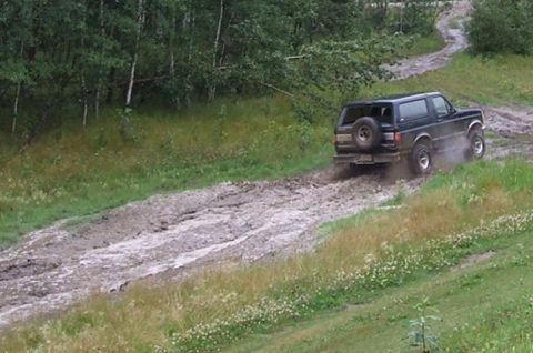 1988 Ford Bronco - A dark-colored Ford Bronco navigates a challenging muddy path, kicking up dirt as it goes.