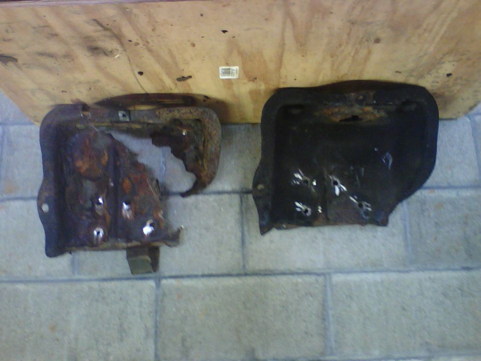 Two stamped steel vehicle parts on pavers: one heavily rusted with holes, one black and intact.