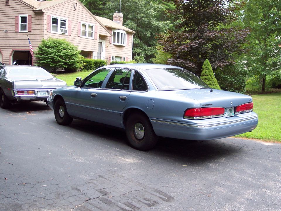 Light blue 2001 Ford Crown Victoria, rear 3/4 view, parked on asphalt driveway next to a house