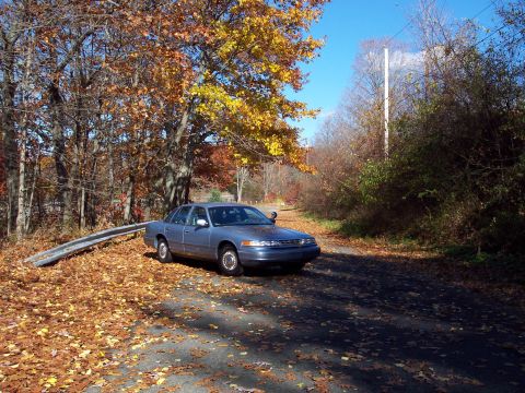 2001 Ford Crown Victoria - A blue 2001 Ford Crown Victoria is parked on a road covered in fallen autumn leaves, with trees displaying fall colors in the background.