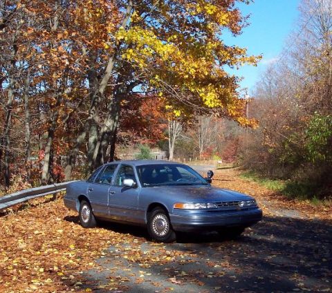 2001 Ford Crown Victoria - A blue 2001 Ford Crown Victoria sits parked on a road blanketed with autumn leaves.